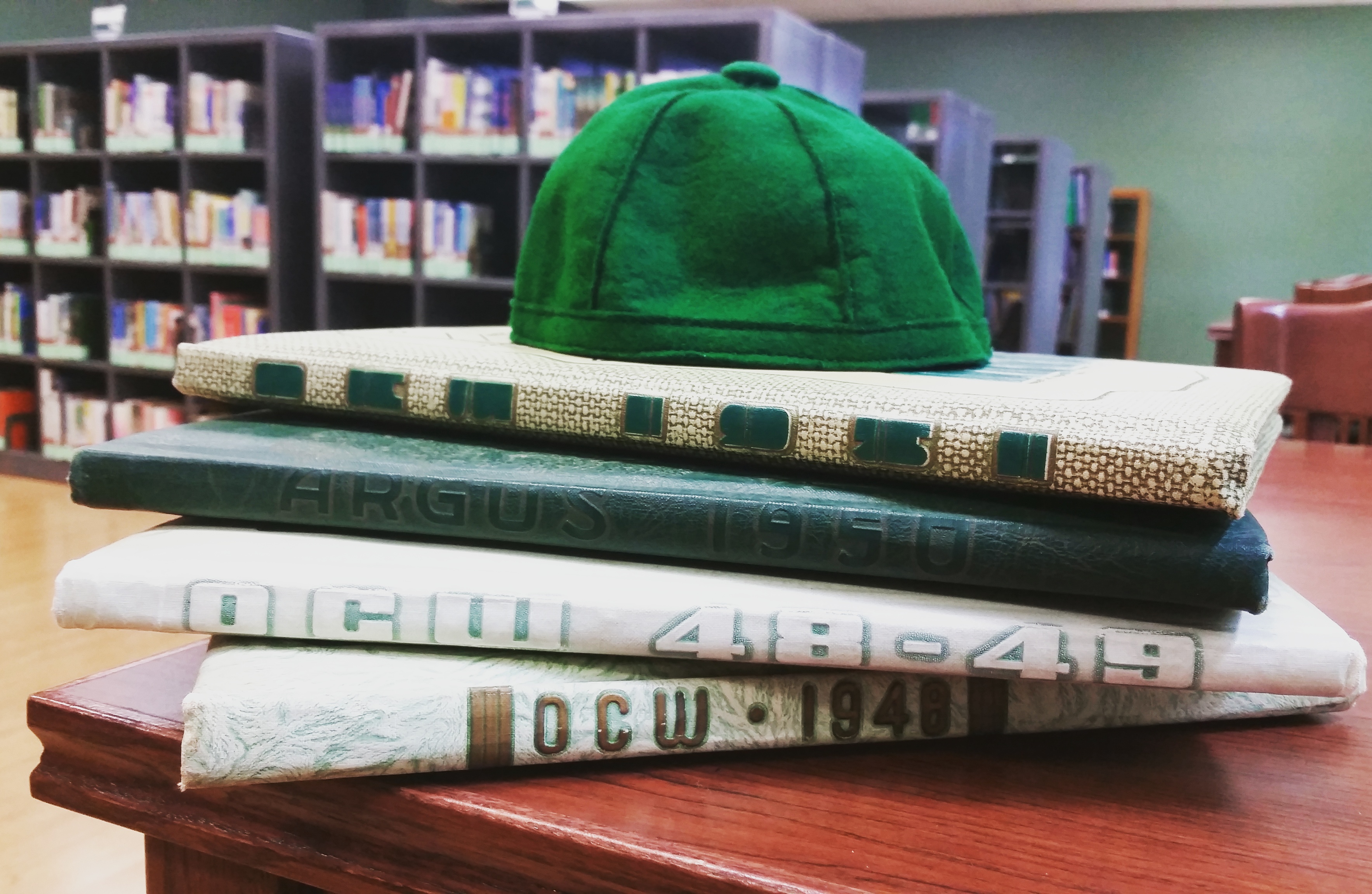 in Nash Library room 301, a green felt beanie atop a stack of four Oklahoma College for Women yearbooks