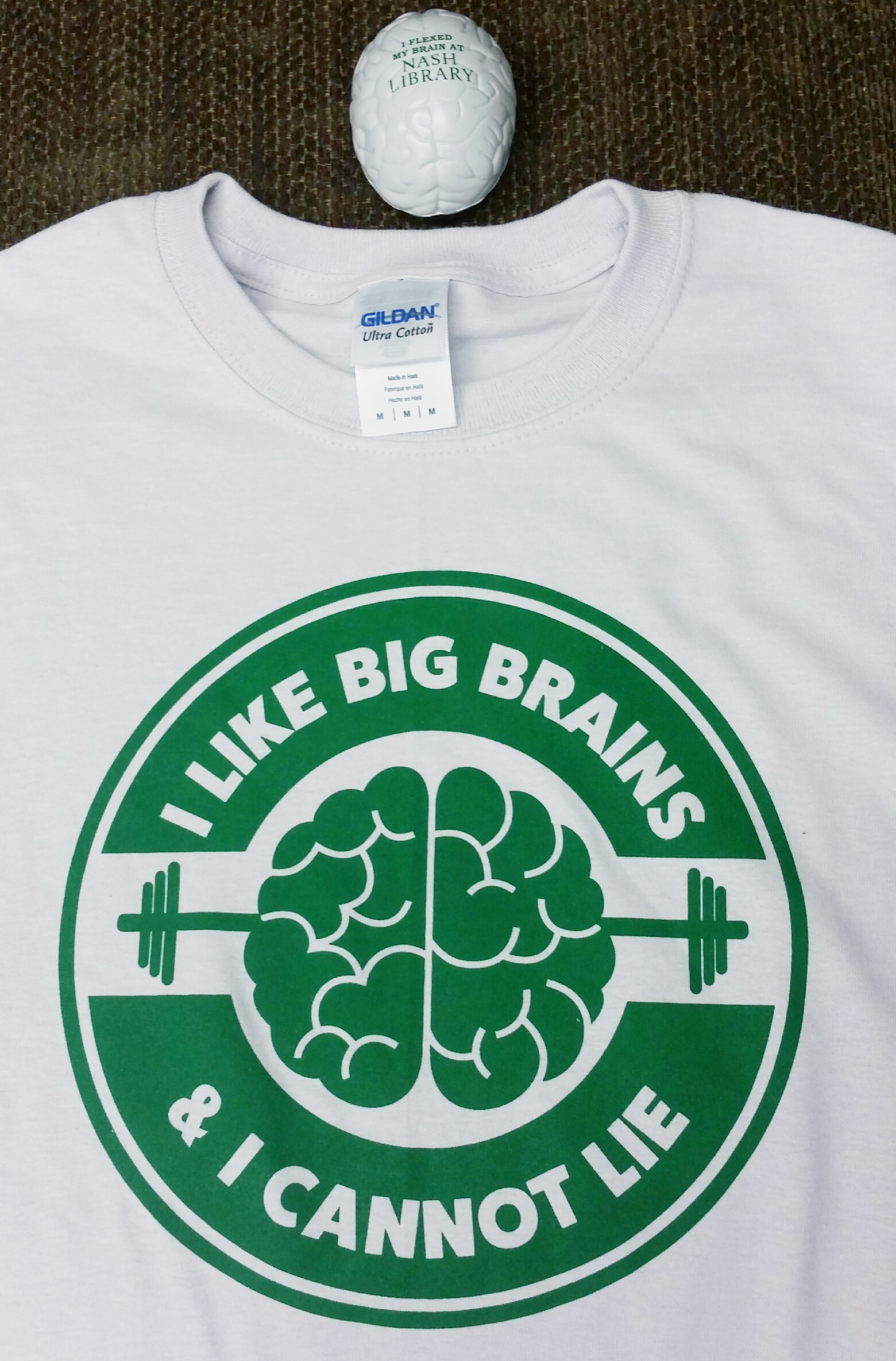 gray t-shirt with green screen printed logo showing a weightlifting brain with text saying I like big brains and I cannot lie. Above the t-shirt, there is a gray squeeze ball shaped like a brain with printed green text on it that says I flexed my brain at Nash Library
